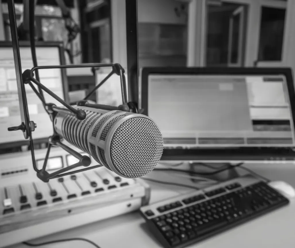 Micro de studio de radio sur son bras articulé, devant une table de mixage et un écran d'ordinateur, en noir et blanc.